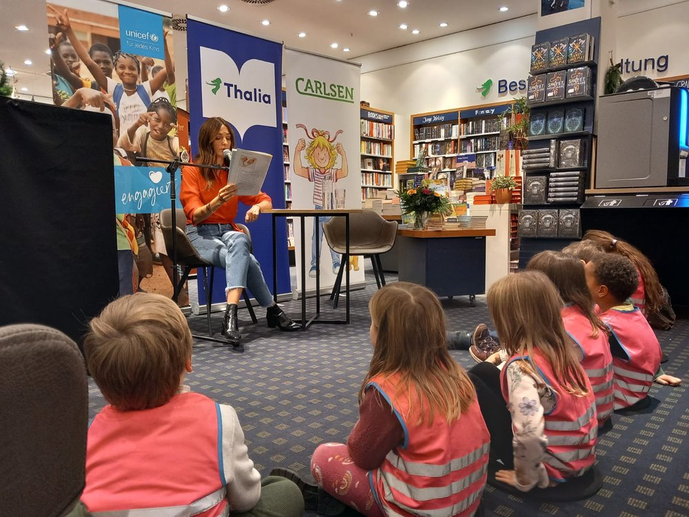 Eine Frau liest in einer Bibliothek aus einem Buch vor. Eine Gruppe von Kindern sitzt auf dem Boden und hört aufmerksam zu.