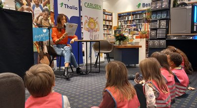 Eine Frau liest in einer Bibliothek aus einem Buch vor. Eine Gruppe von Kindern sitzt auf dem Boden und hört aufmerksam zu.